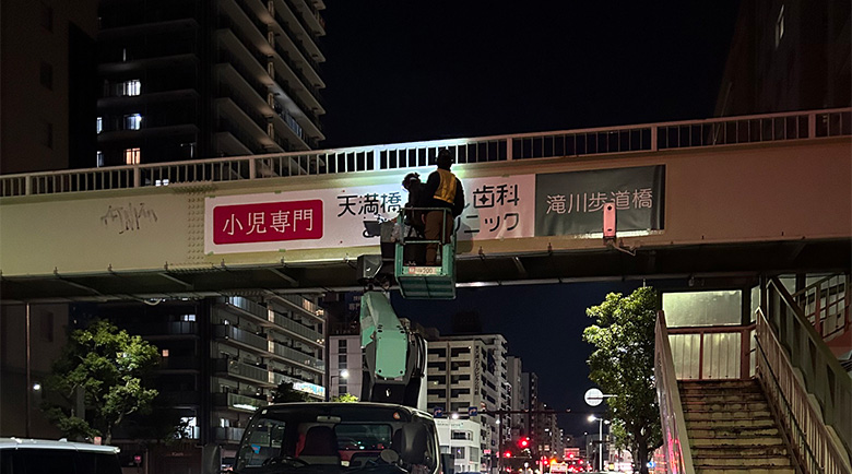 高所作業車に乗って歩道橋へシート施工を行う作業員の遠景